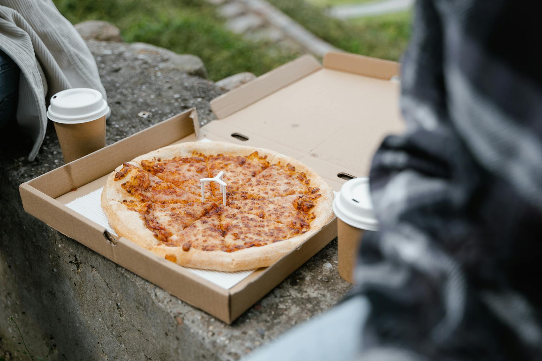 Pizza w pudełku na kamiennej ławce obok kubków kawy na dworze