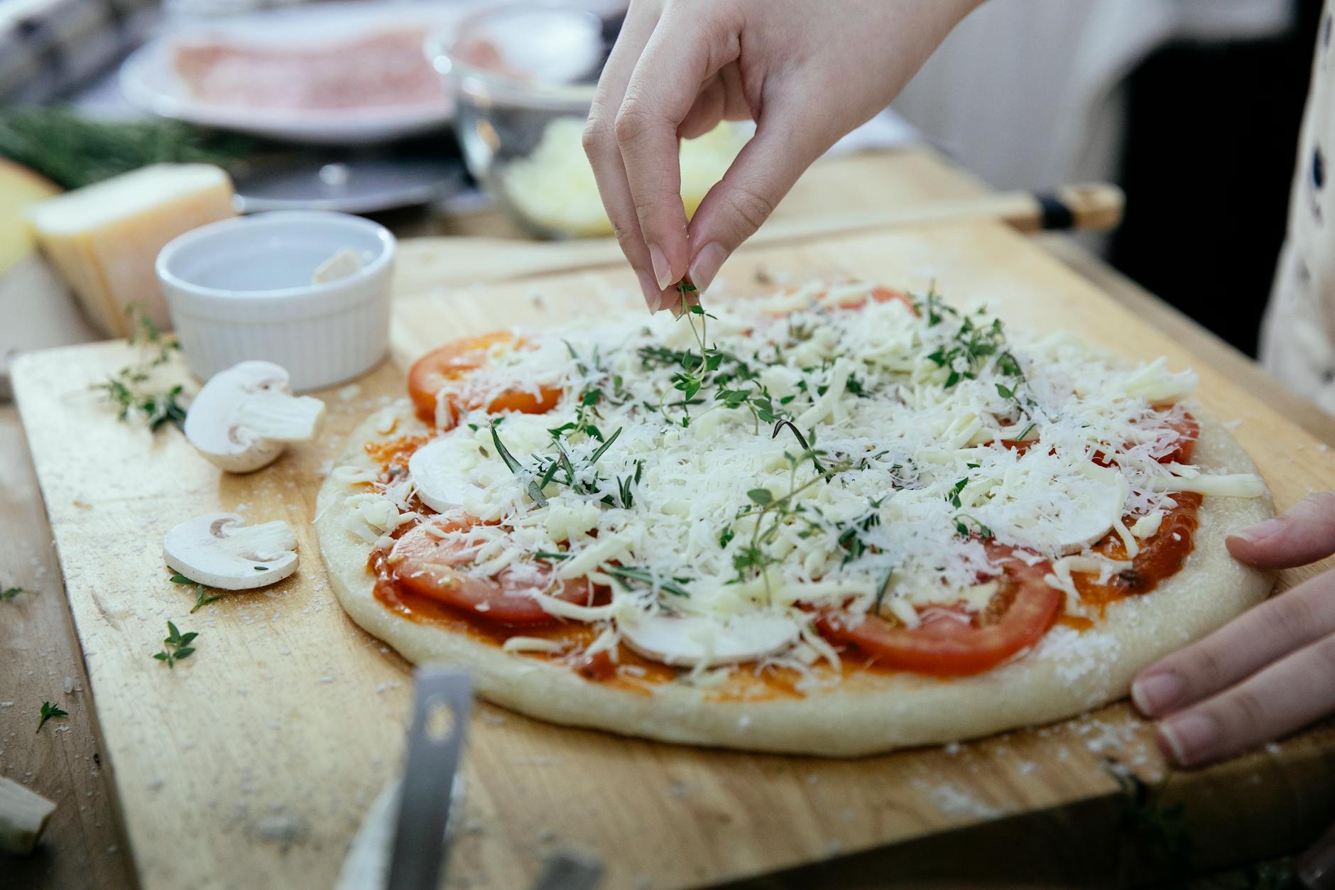 Kobieta układa świeże zioła na surowej pizzy z pomidorami i pieczarkami