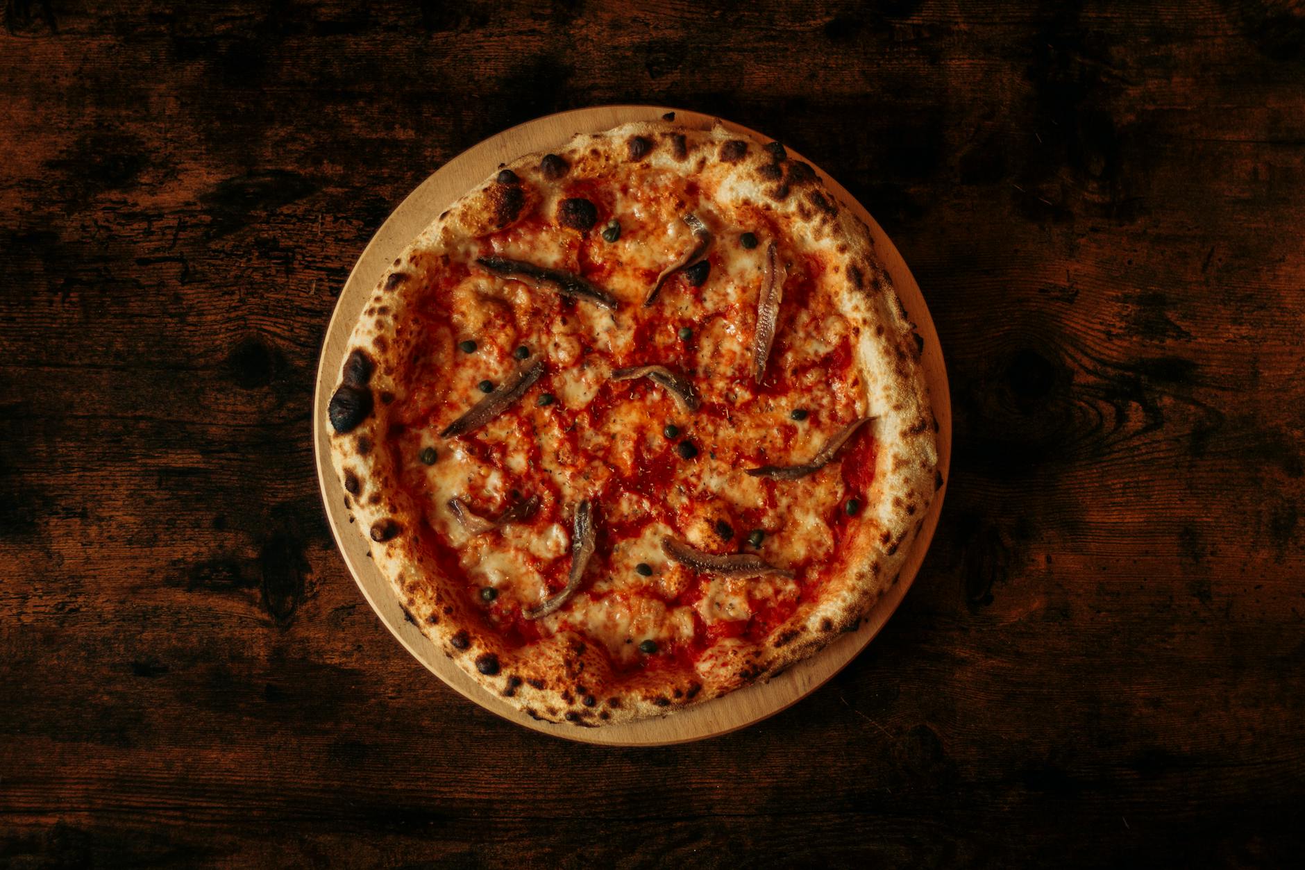 Sos do pizzy z dodatkiem anchois i kaparów wytrawna propozycja dla koneserów Pizza z anchois i sosem pomidorowym na rustykalnym drewnianym stole