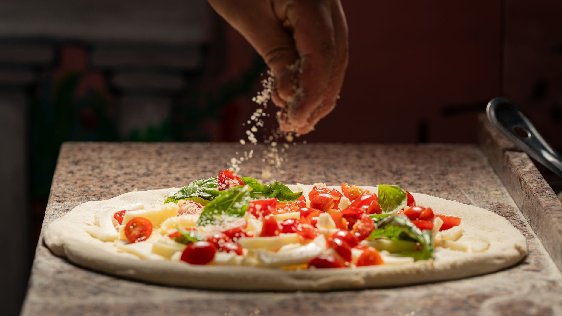Kucharz posypuje pizzę serem obok świeżych pomidorów i bazylii